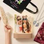 Δώρα για κολλητή σε flat lay σύνθεση: φωτιζόμενο κάδρο με φωτογραφία φίλων, κολιέ φιλίας με καρδιά, παλέτα σκιών Naked3, σατέν ρόμπα και προσωποποιημένη tote bag