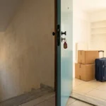 Ανοιχτή πόρτα φοιτητικού σπιτιού με κλειδιά στην κλειδαριά, κούτες και βαλίτσα την ημέρα της φοιτητικής μετακόμισης