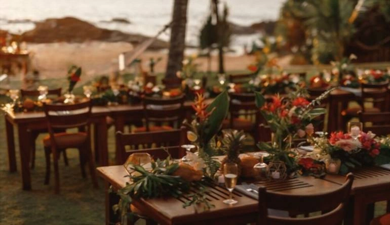 Ξύλινα τραπέζια για δείπνο γάμου διακοσμημένα με τροπικά λουλούδια, ανανάδες, καρύδες και γυάλινα φωτιστικά.