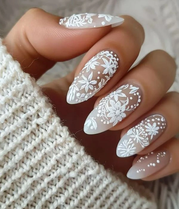 Νυφικά νύχια με λευκά σχέδια δαντέλας και λουλουδιών πάνω σε nude βάση, με αμυγδαλωτό σχήμα.