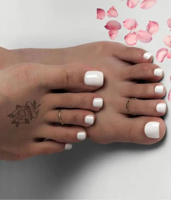Λευκά νυφικά νύχια ποδιών σε τετράγωνο σχήμα, διακοσμημένα με δύο χρυσά δαχτυλίδια και ένα τατουάζ με τριαντάφυλλο στον αστράγαλο, με ροζ πέταλα λουλουδιών στο φόντο.
