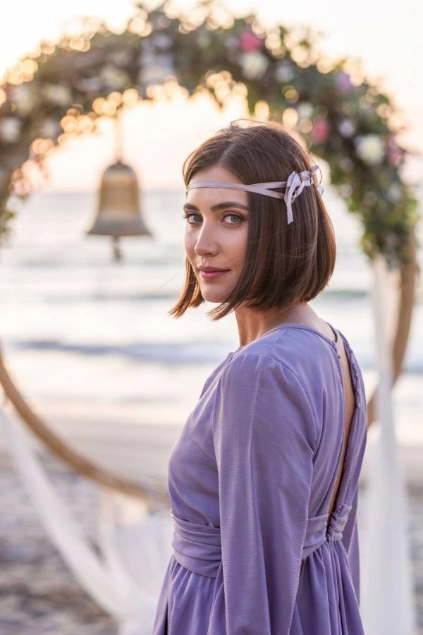 Λείο καρέ με αξεσουάρ κορδέλα για καλεσμένη με κοντά μαλλιά σε γάμο. Η γυναίκα έχει καστανά ίσια μαλλιά και φοράει μια λιλά κορδέλα δεμένη στο πλάι. Φοράει ένα λιλά φόρεμα με ανοιχτή πλάτη και κοιτάζει πάνω από τον ώμο της. Βρίσκεται σε μια παραλία μπροστά από μια διακοσμημένη αψίδα γάμου με λουλούδια και μια καμπάνα. Το ηλιοβασίλεμα δημιουργεί ένα ζεστό φως.