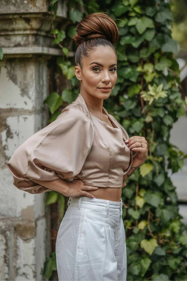 Κομψός ψηλός κότσος για καλεσμένη με κοντά μαλλιά σε γάμο. Η γυναίκα έχει καστανά μαλλιά πιασμένα ψηλά σε έναν λείο κότσο. Φοράει ένα μπεζ σατέν τοπ με φουσκωτά μανίκια και λευκό παντελόνι. Ποζάρει σε εξωτερικό χώρο με έναν τοίχο καλυμμένο με αναρριχώμενα φυτά στο φόντο.