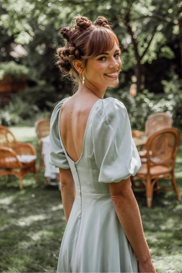 Πρωτότυπο χτένισμα με μικρούς κότσους και αφέλειες για καλεσμένη με κοντά μαλλιά σε γάμο. Η γυναίκα έχει καστανά μαλλιά και χαμογελά κοιτάζοντας πάνω από τον ώμο της. Φοράει ένα ανοιχτό πράσινο φόρεμα με φουσκωτά μανίκια και πέρλες στα αυτιά. Ποζάρει σε έναν κήπο με ψάθινες καρέκλες στο φόντο.