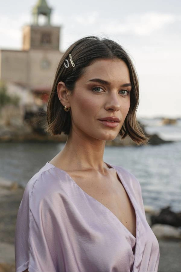 Chic κοντό καρέ χτένισμα για καλεσμένη σε γάμο με χωρίστρα στο πλάι και διακοσμητικά χρυσά τσιμπιδάκια. Η γυναίκα έχει καστανά μαλλιά και φοράει ένα απαλό λιλά σατέν φόρεμα. Ποζάρει σε εξωτερικό χώρο με θάλασσα και ένα παλιό κτίριο στο φόντο.