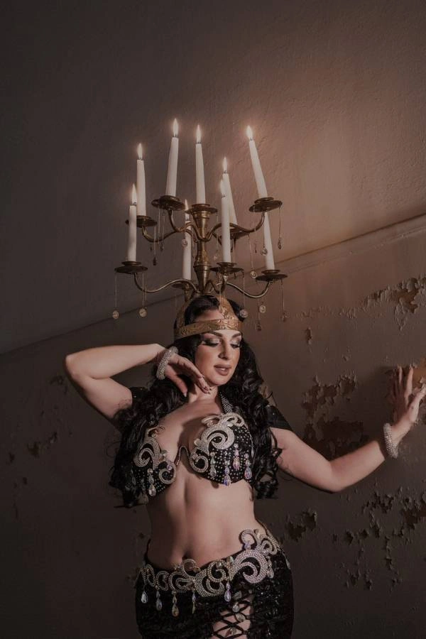 Η Ευδοκία Durrah, χορεύτρια Oriental, ποζάρει σε σκοτεινό, ατμοσφαιρικό φόντο με έναν παλιό τοίχο. Φοράει ένα εντυπωσιακό μαύρο κουστούμι χορού με περίτεχνες ασημί και χρυσές λεπτομέρειες και κεντήματα, ενώ ισορροπεί ένα οκτάφωτο κηροπήγιο με αναμμένα κεριά στο κεφάλι της.