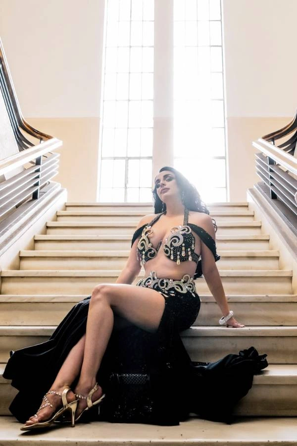 Η Ευδοκία Durrah, χορεύτρια Oriental, κάθεται κομψά σε μια μεγάλη μαρμάρινη σκάλα, φορώντας ένα εντυπωσιακό μαύρο κουστούμι χορού με περίτεχνα ασημί και χρυσά κεντήματα.