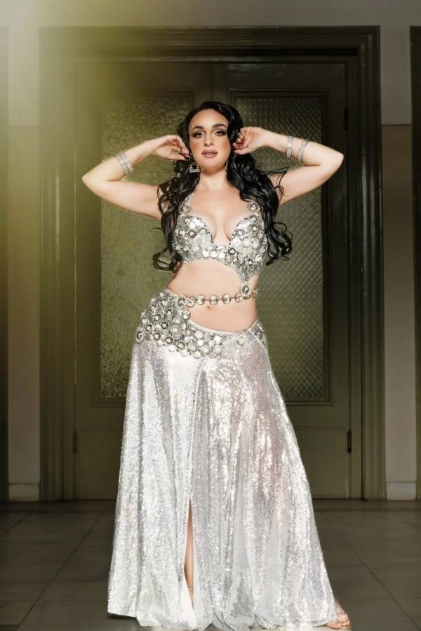 Η Ευδοκία Durrah, καταξιωμένη χορεύτρια Oriental, ποζάρει όρθια μπροστά από μια ξύλινη πόρτα, φορώντας ένα αστραφτερό, ασημί κουστούμι χορού με καθρέφτες και στρας.