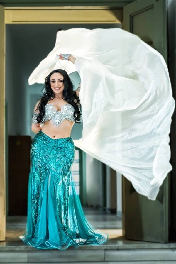 Η Ευδοκία Durrah, καταξιωμένη χορεύτρια Oriental, ποζάρει χαμογελαστή και δυναμική σε μια πόρτα, φορώντας ένα ασημί τοπ χορού και μια εντυπωσιακή μπλε-τιρκουάζ φούστα με πούλιες και κεντήματα.