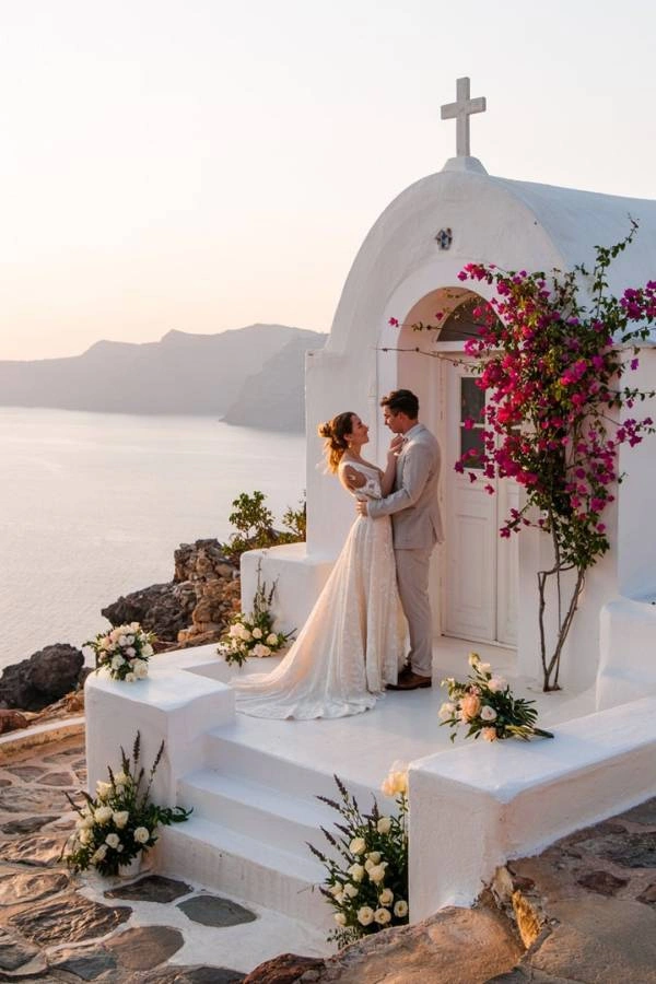 Ένα ζευγάρι αγκαλιά μπροστά σε ένα λευκό ξωκλήσι στολισμένο με λουλούδια, με θέα στη θάλασσα και τα βουνά κατά τη δύση του ηλίου, αποτυπώνοντας τη ρομαντική ατμόσφαιρα ενός γάμου σε νησί.