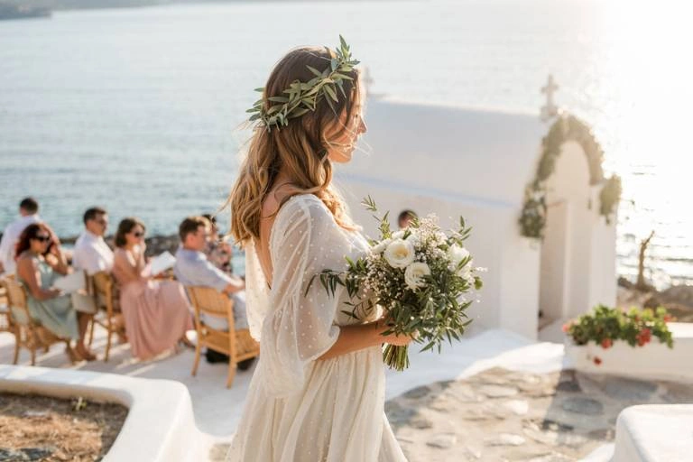 Ονειρεμένος γάμος σε νησί. Νύφη με στεφάνι ελιάς και νυφική ανθοδέσμη περπατά προς ένα λευκό ξωκλήσι δίπλα στη θάλασσα, με καλεσμένους να παρακολουθούν.