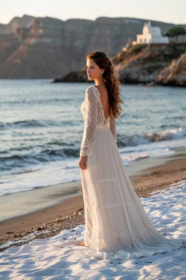 Μια νύφη με ένα αέρινο νυφικό στέκεται στην αμμουδιά καθώς δύει ο ήλιος, κοιτώντας τη θάλασσα, με ένα γραφικό ξωκλήσι να διακρίνεται στο λόφο στο βάθος, εικόνα που αποτυπώνει την ομορφιά ενός νησιώτικου γάμου.