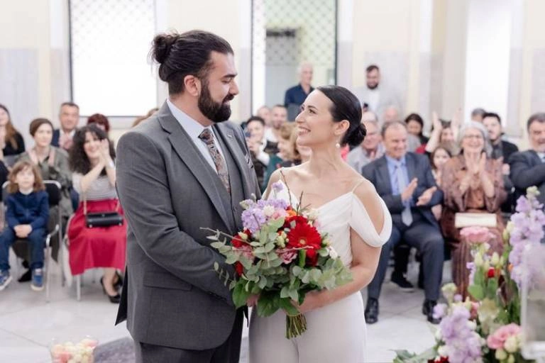 Νύφη και γαμπρός με χαμόγελο κατά τη διάρκεια της τελετής του πολιτικού γάμου, μπροστά σε φίλους και συγγενείς.