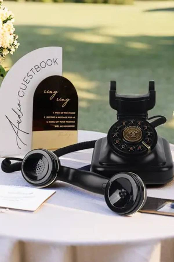 Διακόσμηση γάμου με μαύρο ρετρό τηλέφωνο σε τραπέζι ως audio guest book, πλαισιωμένο από λουλούδια και πίνακα οδηγιών