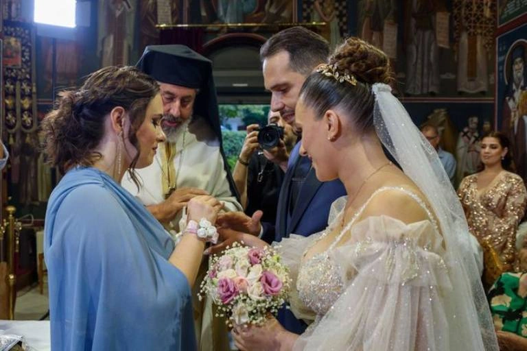 Στιγμιότυπο από γάμο όπου η κουμπάρα βάζει τη βέρα στον γαμπρό μπροστά στον ιερέα και τη νύφη