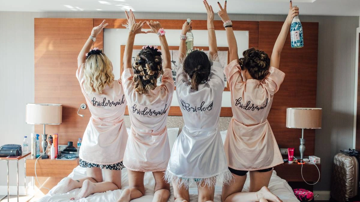 Η νύφη με τις φίλες της φοράνε σατέν ρόμπες και γιορτάζουν το bachelorette party μέσα σε δωμάτιο ξενοδοχείου, γεμάτο χαρά και ενθουσιασμό.