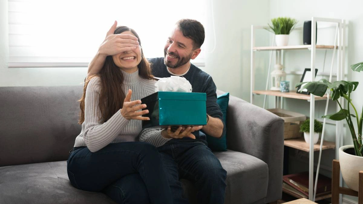 Χαμογελαστό ζευγάρι καθισμένο σε καναπέ, με τον άντρα να καλύπτει τα μάτια της γυναίκας και να της προσφέρει δώρο σε μπλε κουτί, σε ρομαντική στιγμή για επέτειο.