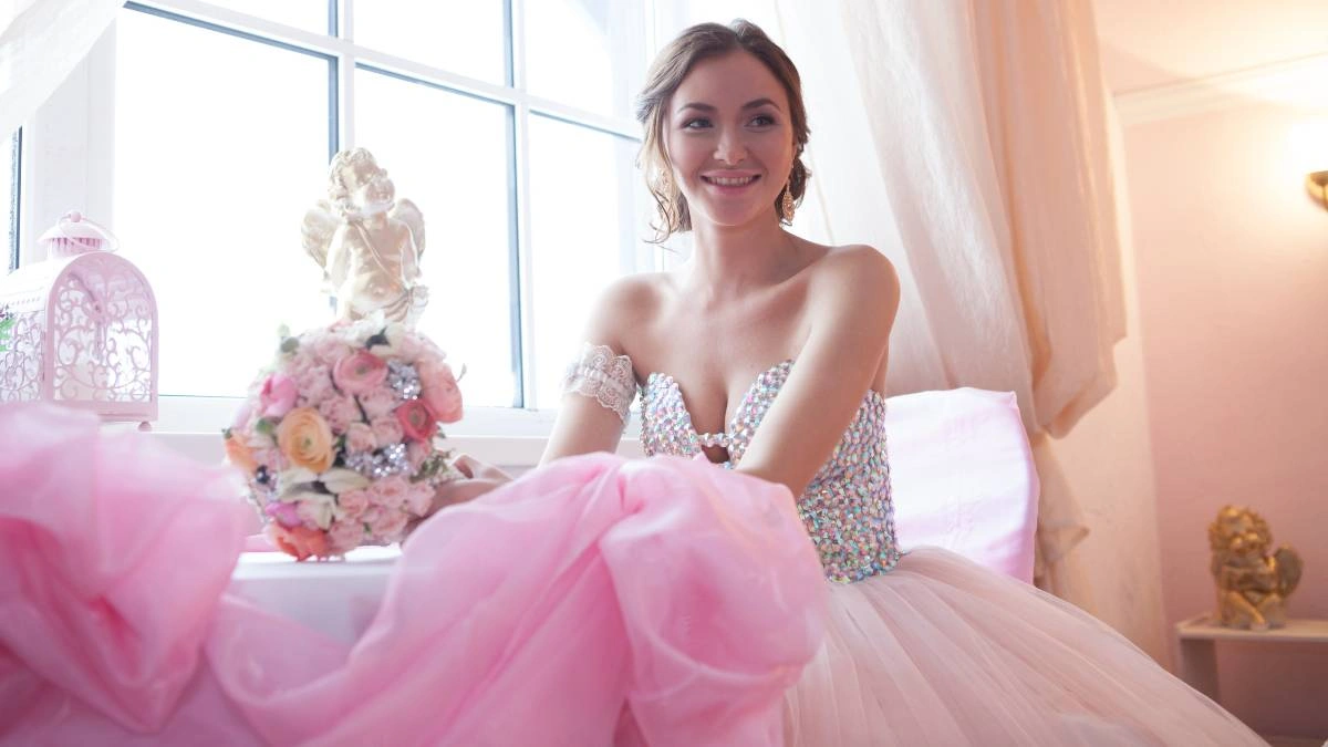 Νύφη με ρομαντικό φόρεμα και ροζ τούλινη φούστα, κάθεται χαμογελαστή δίπλα σε ανθοδέσμη με παστέλ λουλούδια, δίπλα σε παράθυρο.
