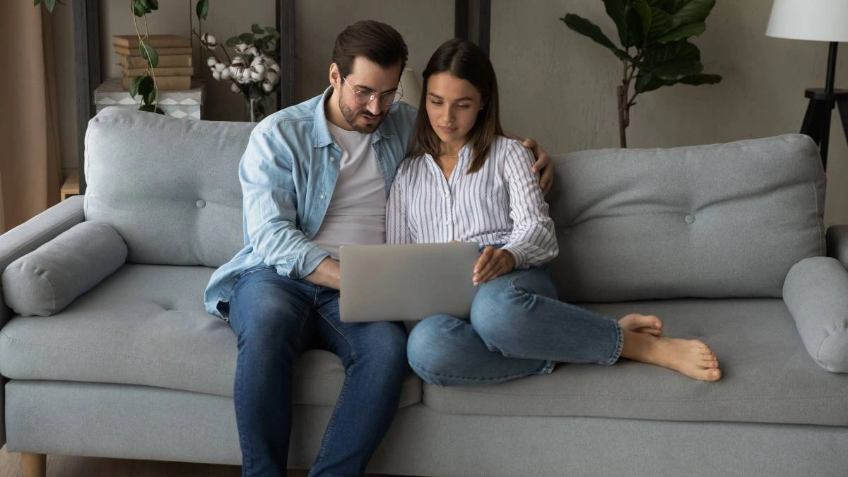 Ζευγάρι κάθεται στον καναπέ και οργανώνει τον γάμο του κοιτάζοντας μαζί ένα λάπτοπ, σε χαλαρή και οικεία ατμόσφαιρα στο σπίτι.