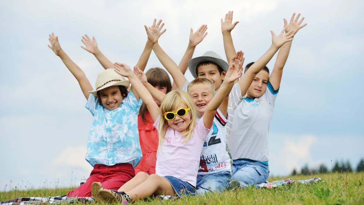 Παιδιά καθισμένα σε γρασίδι με σηκωμένα χέρια και χαμόγελα, απολαμβάνοντας τη φύση σε ηλιόλουστη ημέρα.