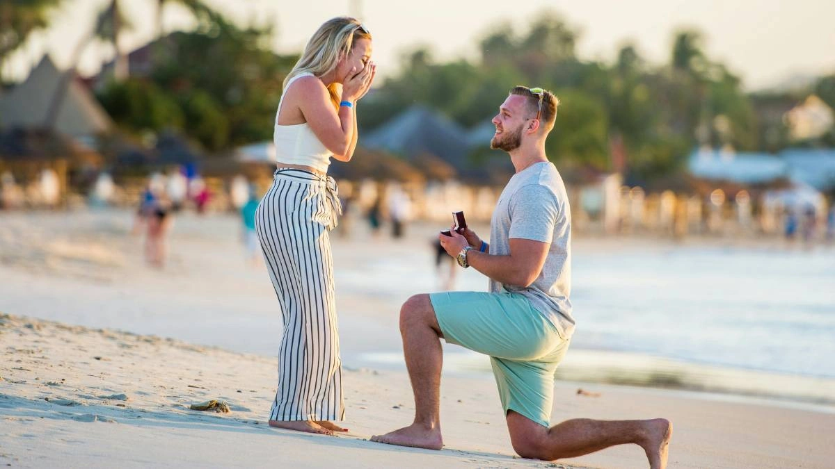 Άντρας γονατιστός στην παραλία κάνει πρόταση γάμου σε γυναίκα που καλύπτει συγκινημένη το πρόσωπό της, με φόντο το ηλιοβασίλεμα και τη θάλασσα.