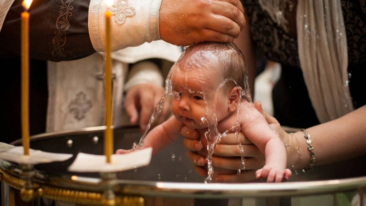 Μωρό την ώρα που βαπτίζεται σε ορθόδοξη τελετή, με ιερέα να ρίχνει νερό στο κεφάλι του και κεριά γύρω από την κολυμπήθρα.