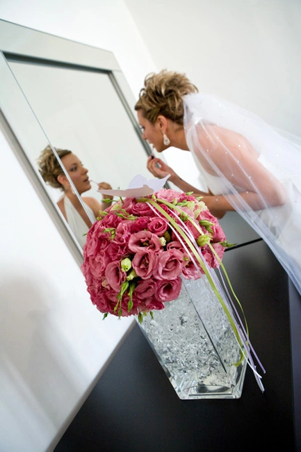 Νύφη μπροστά στον καθρέφτη με ρομαντικό μπουκέτο από ροζ λουλούδια σε γυάλινο βάζο, ετοιμασία πριν τον γάμο από τη Vassiliki Creations