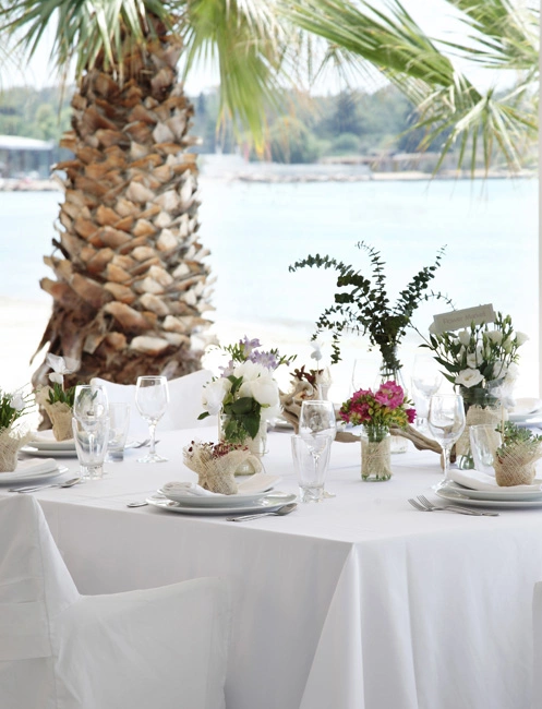 Καλοκαιρινός στολισμός τραπεζιού γάμου σε παραλία με φυσικά λουλούδια, λευκό τραπεζομάντιλο και θέα στη θάλασσα, από τη Vassiliki Creations στο Balux