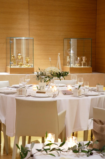 Κομψός στολισμός τραπεζιού γάμου με λευκά λουλούδια και ελιά σε εσωτερικό χώρο δεξίωσης με εκθέματα τέχνης, από τη Vassiliki Creations στο Milos Hilton