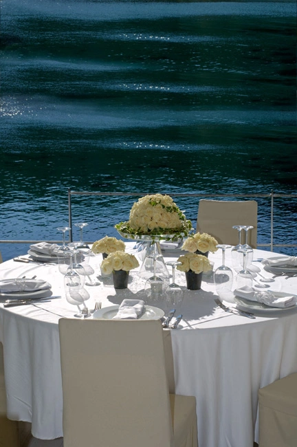 Λευκό τραπέζι γάμου σε παραθαλάσσιο χώρο με φόντο τη θάλασσα και elegant ανθοσυνθέσεις από λευκά τριαντάφυλλα, από τη Vassiliki Creations στο Moorings