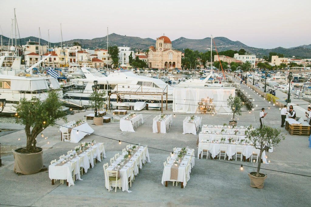 Καλοκαιρινή δεξίωση γάμου με θέα τη μαρίνα της Αίγινας, rustic τραπέζια με λευκά τραπεζομάντιλα και φωτισμός string lights, από τη Vassiliki Creations στον Ναυτικό Όμιλο Αίγινας