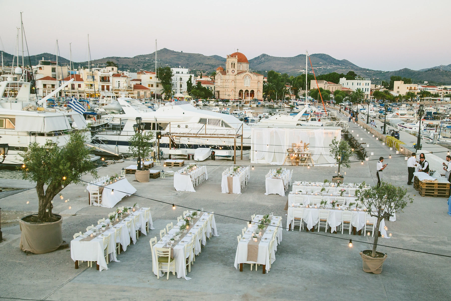 Καλοκαιρινή δεξίωση γάμου με θέα τη μαρίνα της Αίγινας, rustic τραπέζια με λευκά τραπεζομάντιλα και φωτισμός string lights, από τη Vassiliki Creations στον Ναυτικό Όμιλο Αίγινας