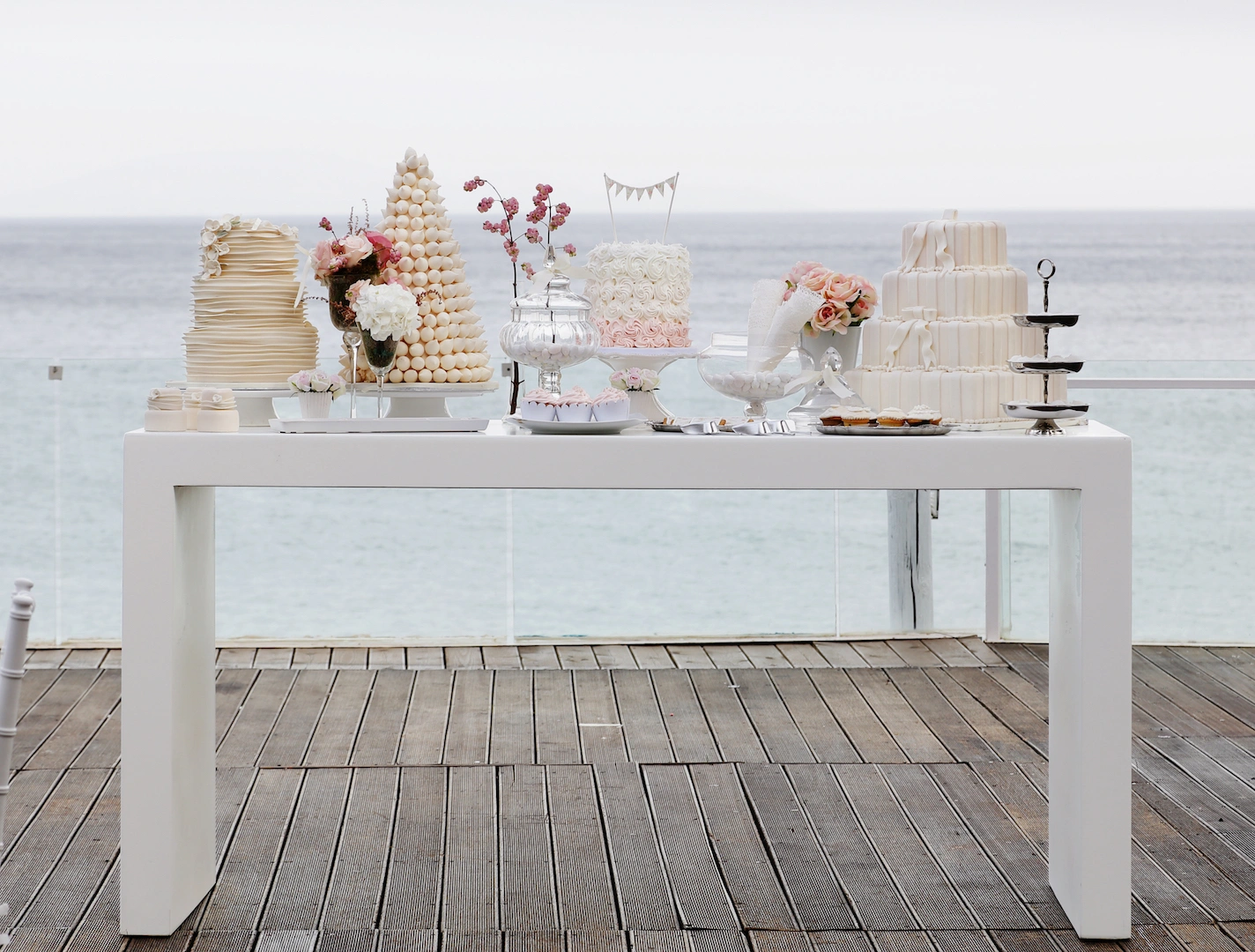 Εντυπωσιακό τραπέζι γλυκών γάμου με θαλασσινό φόντο, τούρτες και ρομαντικά λουλούδια από τη Vassiliki Creations