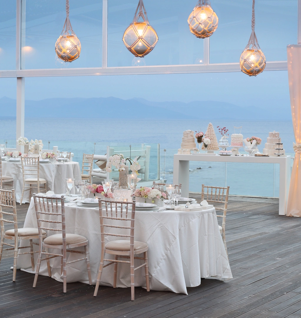 Ρομαντικό σκηνικό δεξίωσης δίπλα στη θάλασσα με λευκά τραπέζια, ροζ λουλούδια και φωτιστικά ρομαντικού στυλ από τη Vassiliki Creations
