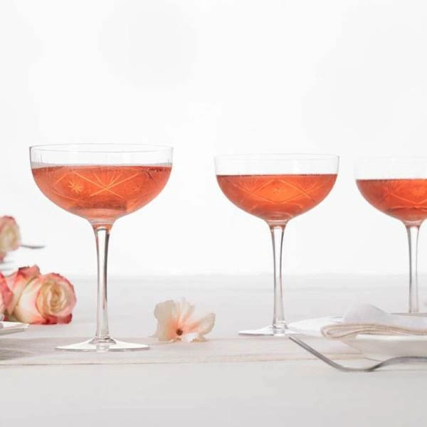 Τρία διάφανα ποτήρια σαμπάνιας τύπου coupe γεμισμένα με ροζ ποτό πάνω σε λευκή επιφάνεια με λουλούδια.
        loading=