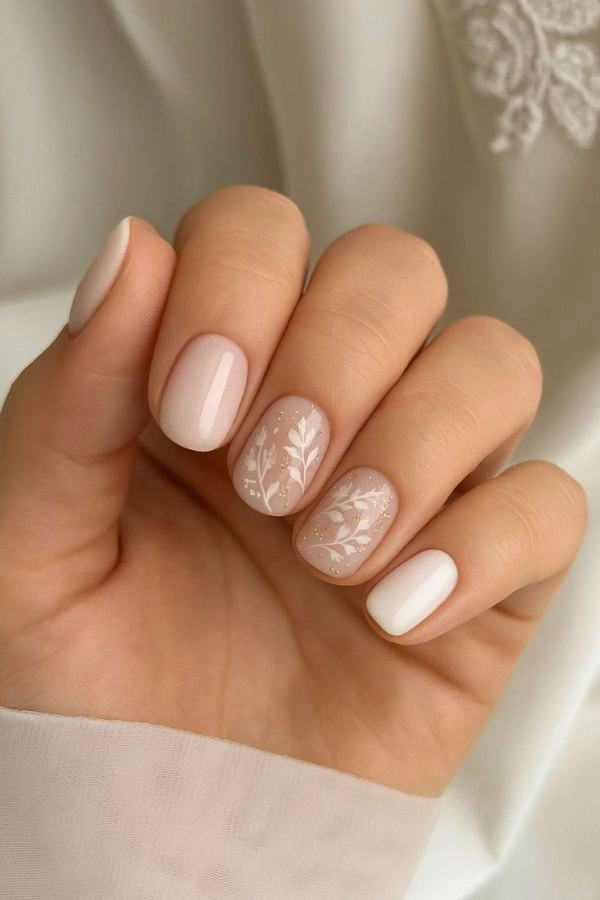 Κοντινό σε νύχια καλεσμένης για γάμο με απαλό ροζ μανικιούρ και λευκό φυτικό σχέδιο σε δύο νύχια, σε φυσικό φως και ουδέτερο φόντο.
