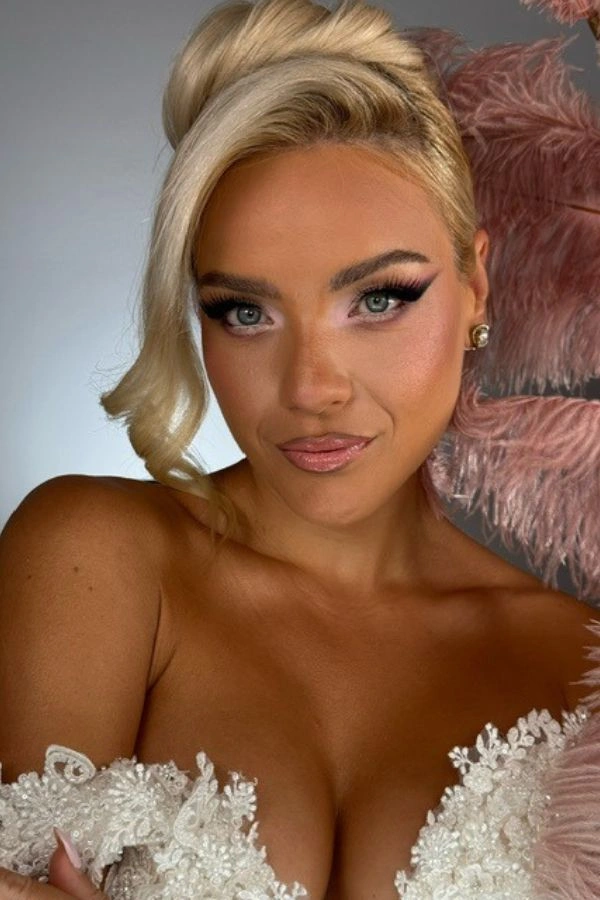 Νυφικό μακιγιάζ με έντονο cat eye eyeliner, λαμπερή βάση και ροζ αποχρώσεις, σε συνδυασμό με κομψό ψηλό σινιόν και ξανθά μαλλιά για glam bridal εμφάνιση.