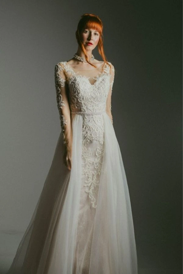 Νυφικό φόρεμα με δαντέλα και διαφάνεια στα μανίκια, ρομαντική γραμμή και αέρινη φούστα, σε minimal και καλλιτεχνικό bridal look.