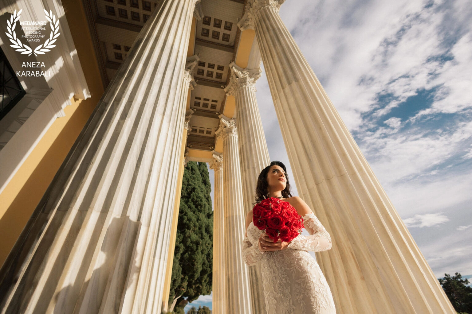 Νύφη με κόκκινη ανθοδέσμη μπροστά σε επιβλητικούς κίονες