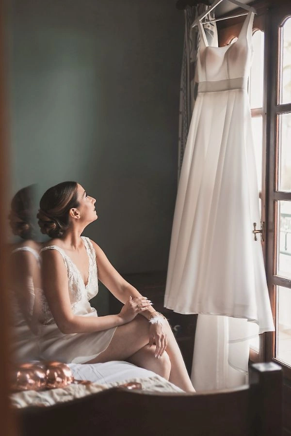 Νύφη στην προετοιμασία του γάμου κοιτά το νυφικό της δίπλα στο παράθυρο