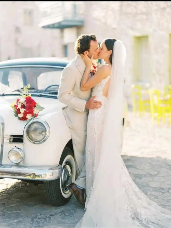 Νεόνυμφο ζευγάρι μπροστά από κλασικό αυτοκίνητο γάμου σε παραδοσιακό χωριό