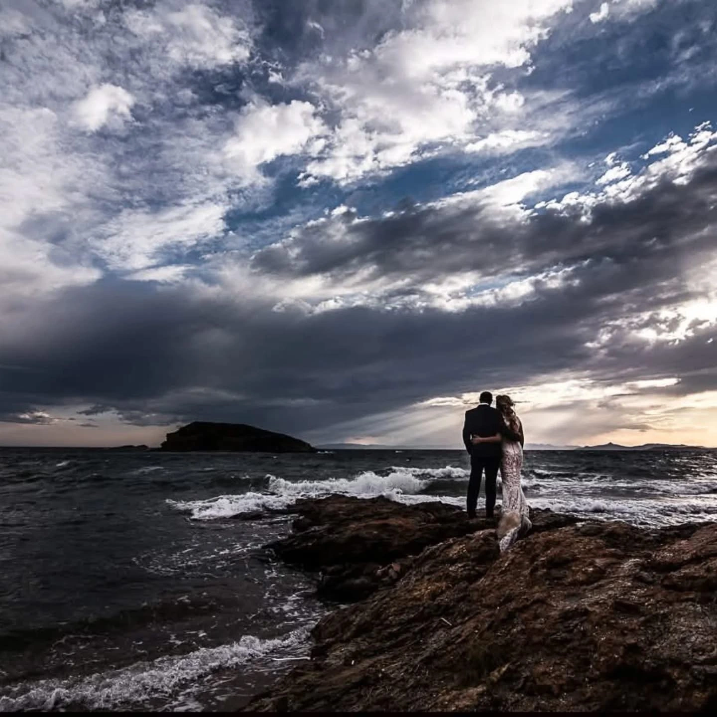 Ζευγάρι αγκαλιά σε βράχια δίπλα στη θάλασσα με δραματικό ουρανό από την Wedding Stars
